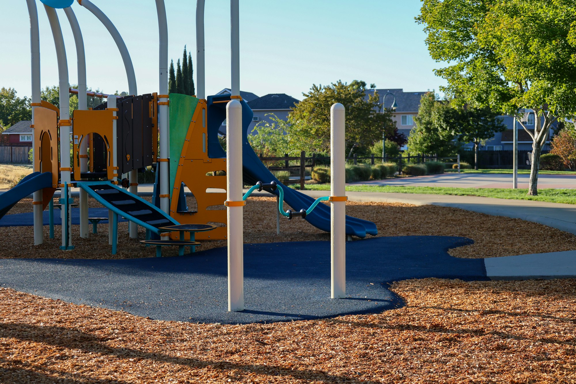Playground with slides, climbing structures, and safety surfacing, surrounded by trees and houses.
