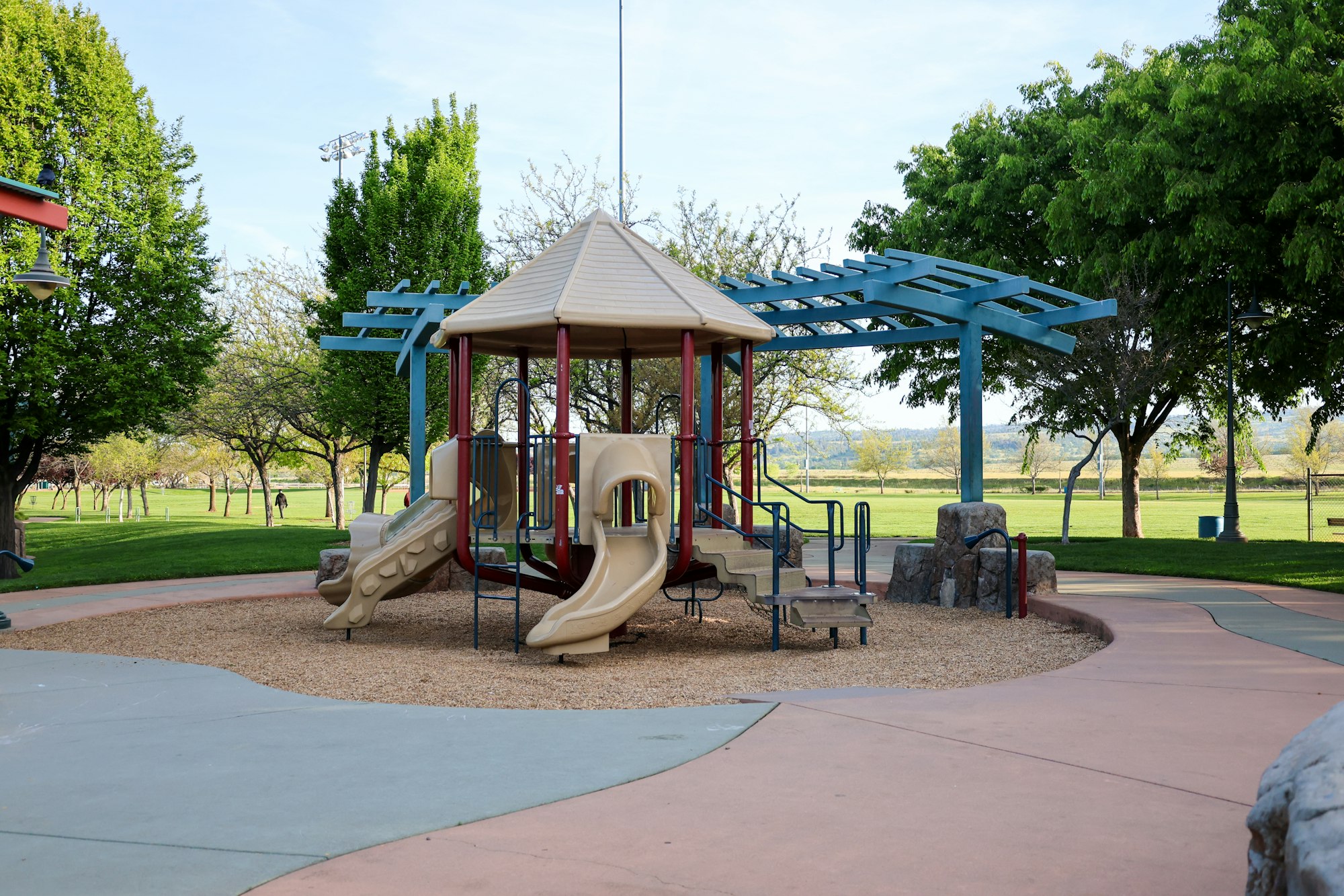 A children's playground with slides and climbing structures, surrounded by green trees and grass.