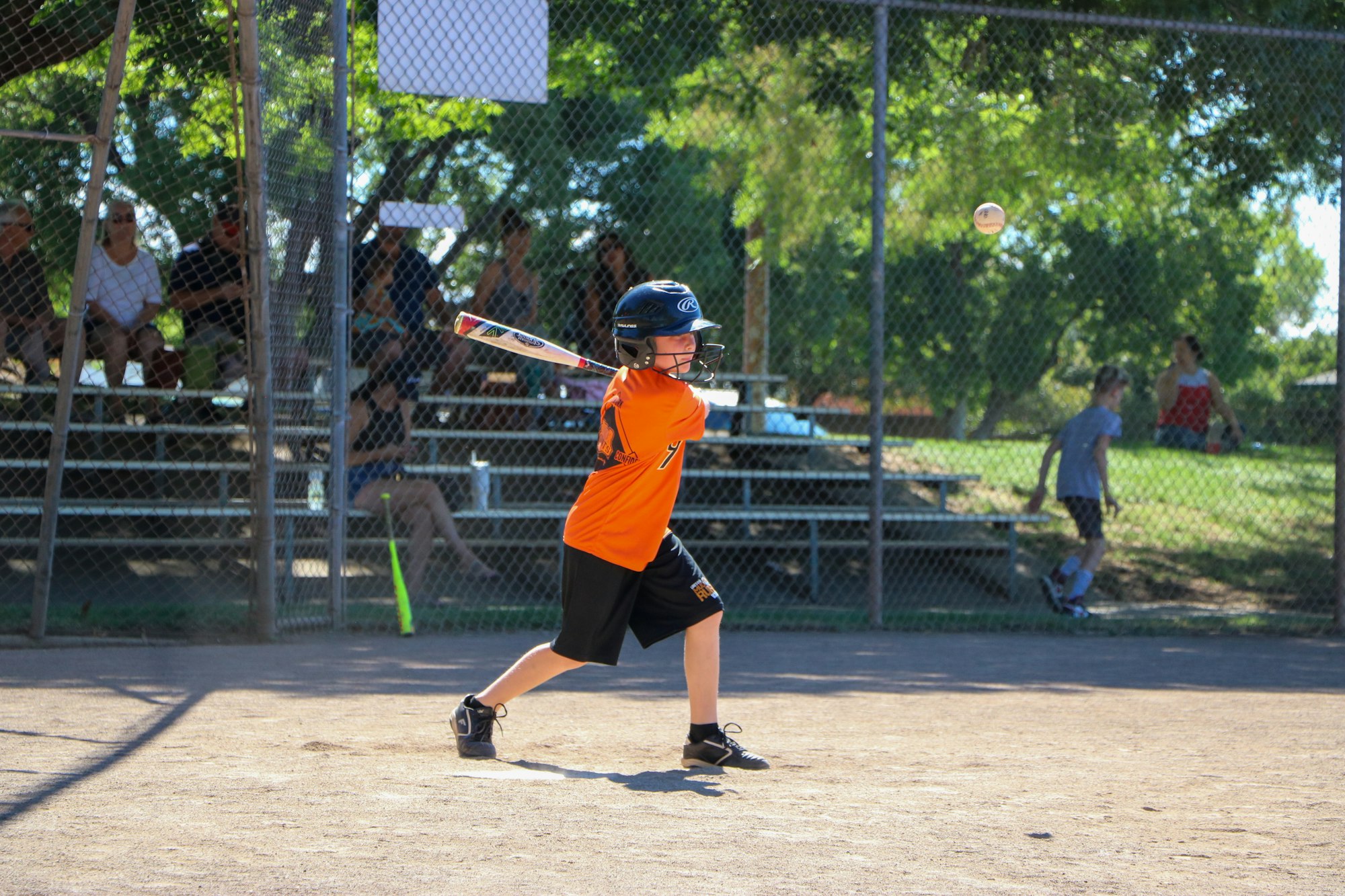 A child in an orange shirt swings a baseball bat on a sunny day, with spectators in the background.