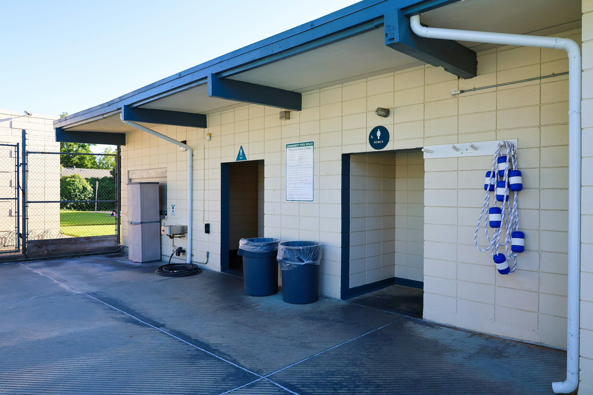 Outdoor changing area with men's and women's signs, trash bins, a coiled hose, and pool rope floats.