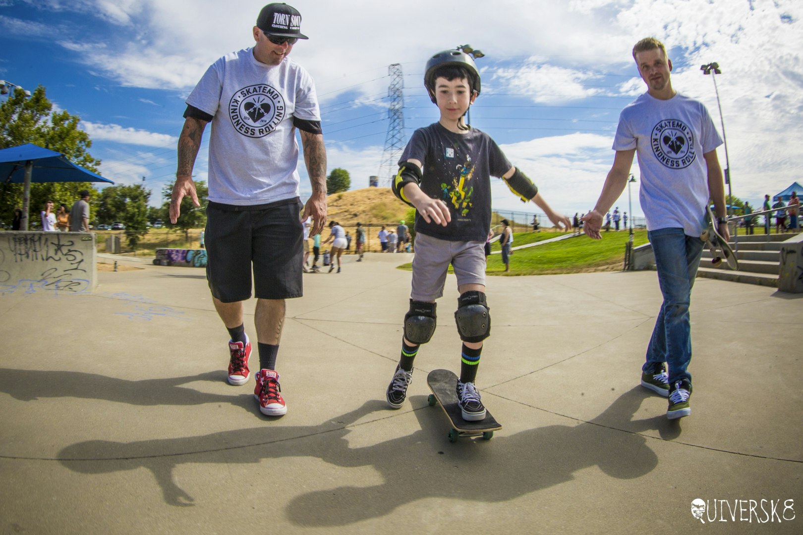 Three people skateboarding, two giving hands to a child learning. Sunny park setting.