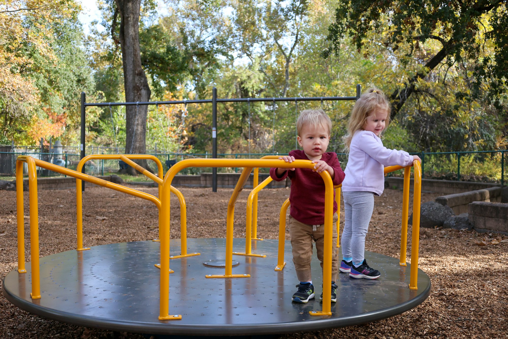 Two children playing on a merry-go-round at a park with trees in the background.