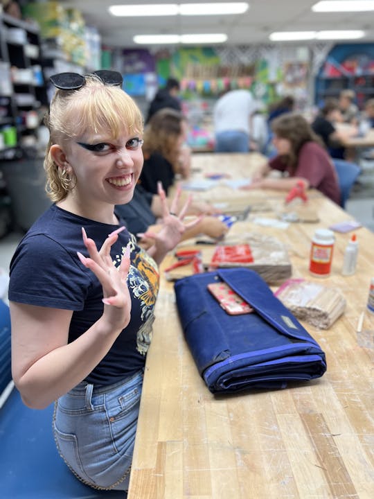A cheerful young woman with unique eye makeup poses at a crafting table, surrounded by others engaged in creative activities.