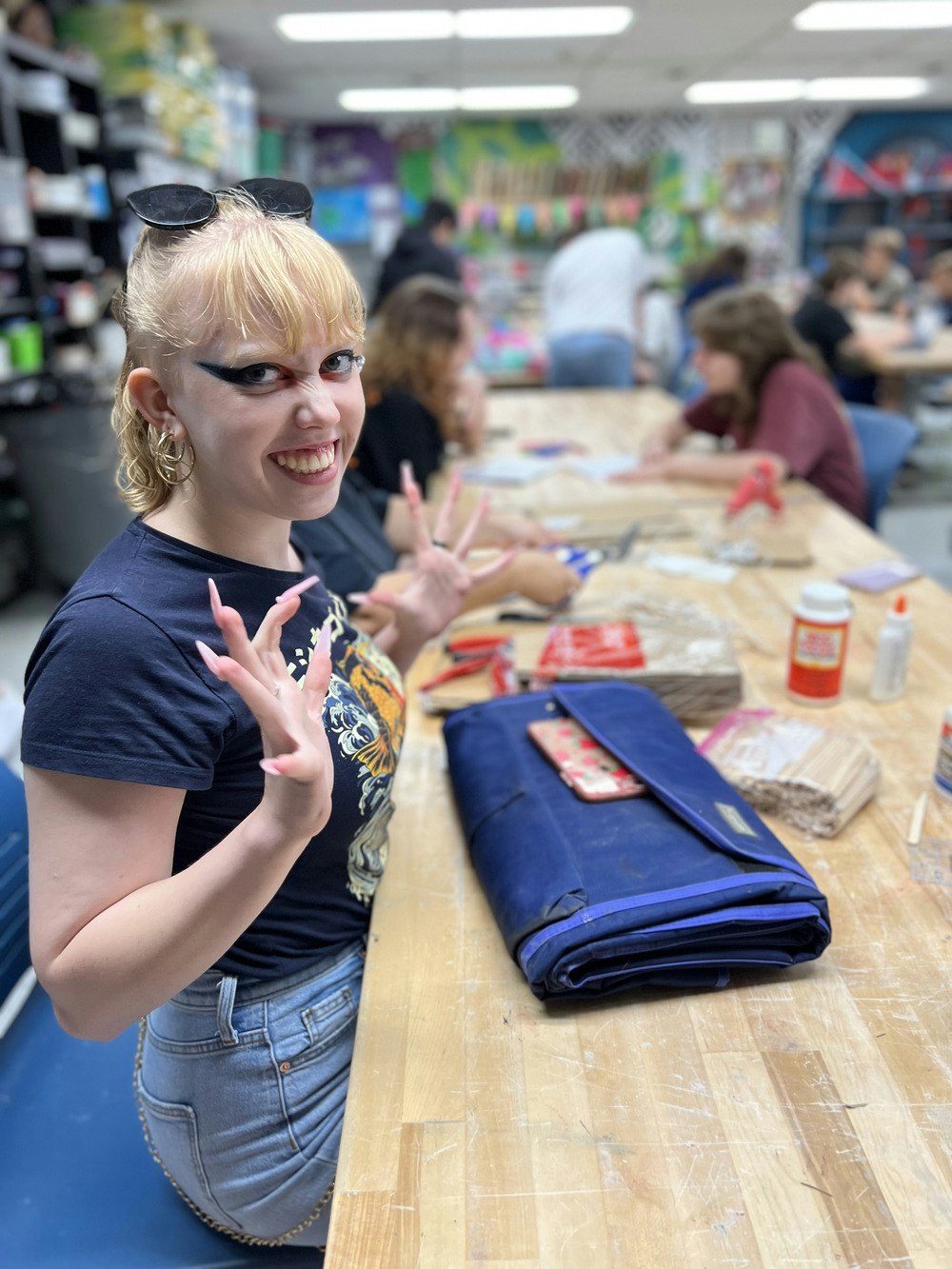 A cheerful young woman with unique eye makeup poses at a crafting table, surrounded by others engaged in creative activities.