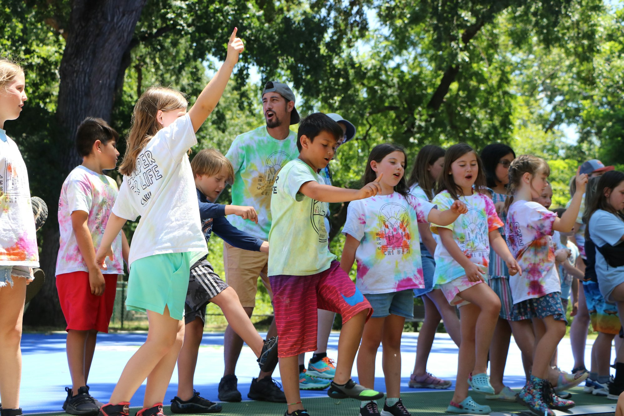 Children and an adult are dancing together outdoors, wearing colorful tie-dye shirts.