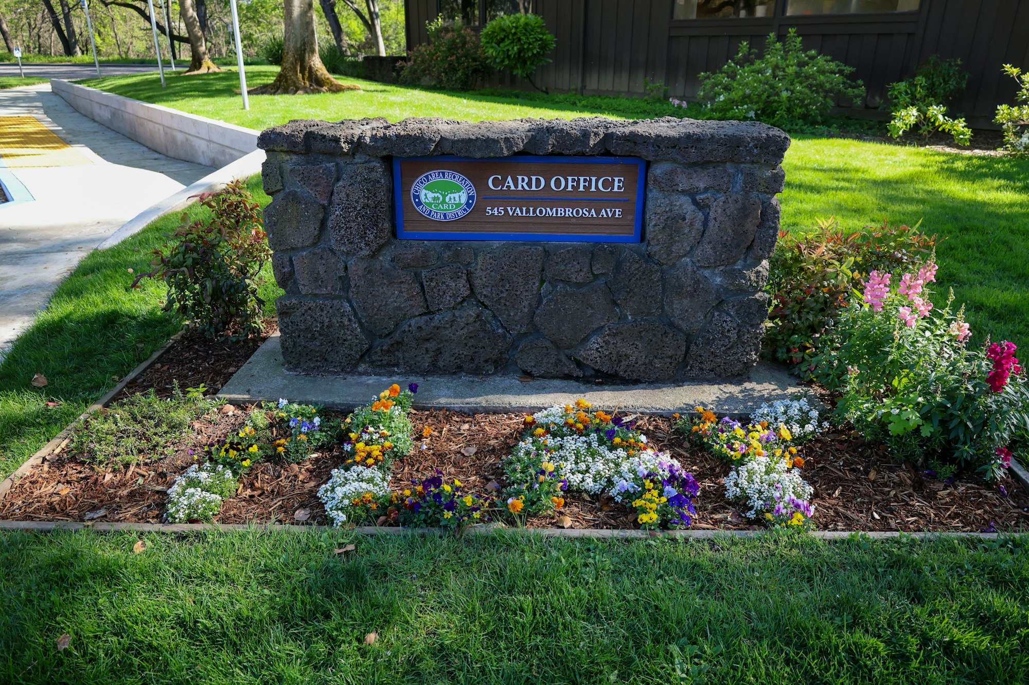 Stone sign for "CARD Office, 545 Vallombrosa Ave" surrounded by colorful flowers and greenery.