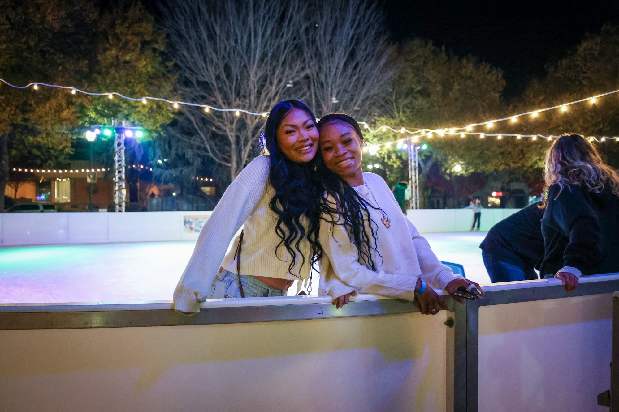 Two people smile at an ice rink at night, surrounded by festive lights and trees.
