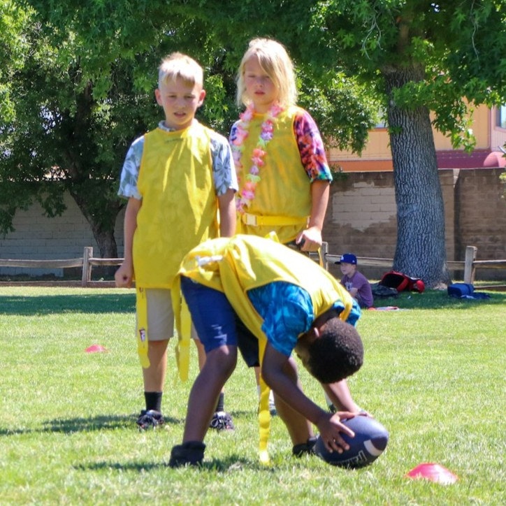 Kids playing on a grassy field, wearing yellow flags, with one child preparing to throw a football while others watch.