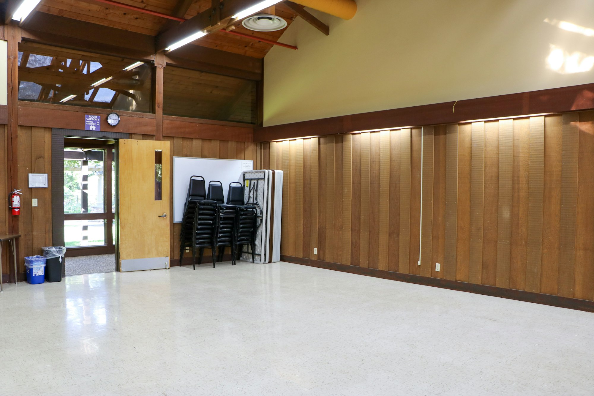 Empty room with stacked chairs, wooden walls, a whiteboard, and a door with glass windows.
