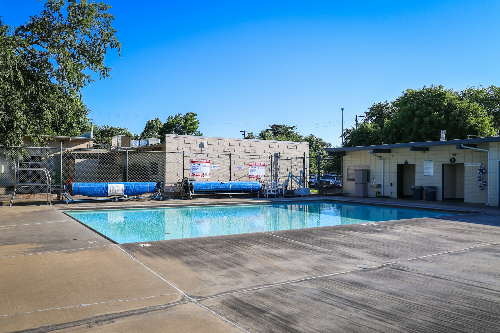 An outdoor swimming pool surrounded by buildings, a fence, and trees on a clear day.