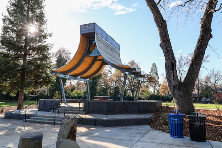 A park with a unique wooden curved structure, trees, benches, trash bins on a sunny day.