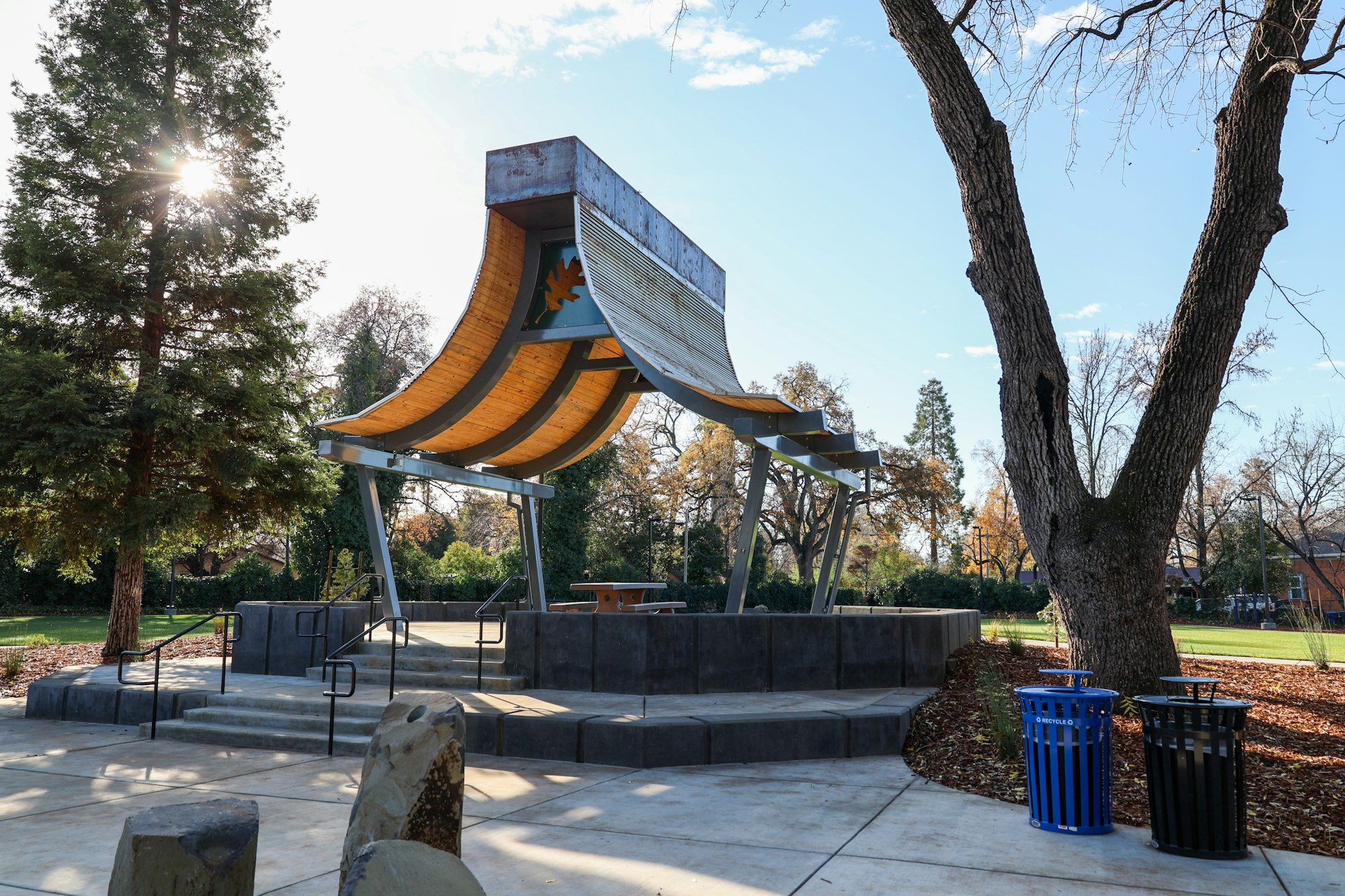 A park with a unique wooden curved structure, trees, benches, trash bins on a sunny day.
