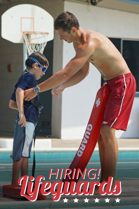 A lifeguard assists a young boy in a pool setting, with a sign promoting lifeguard hiring.