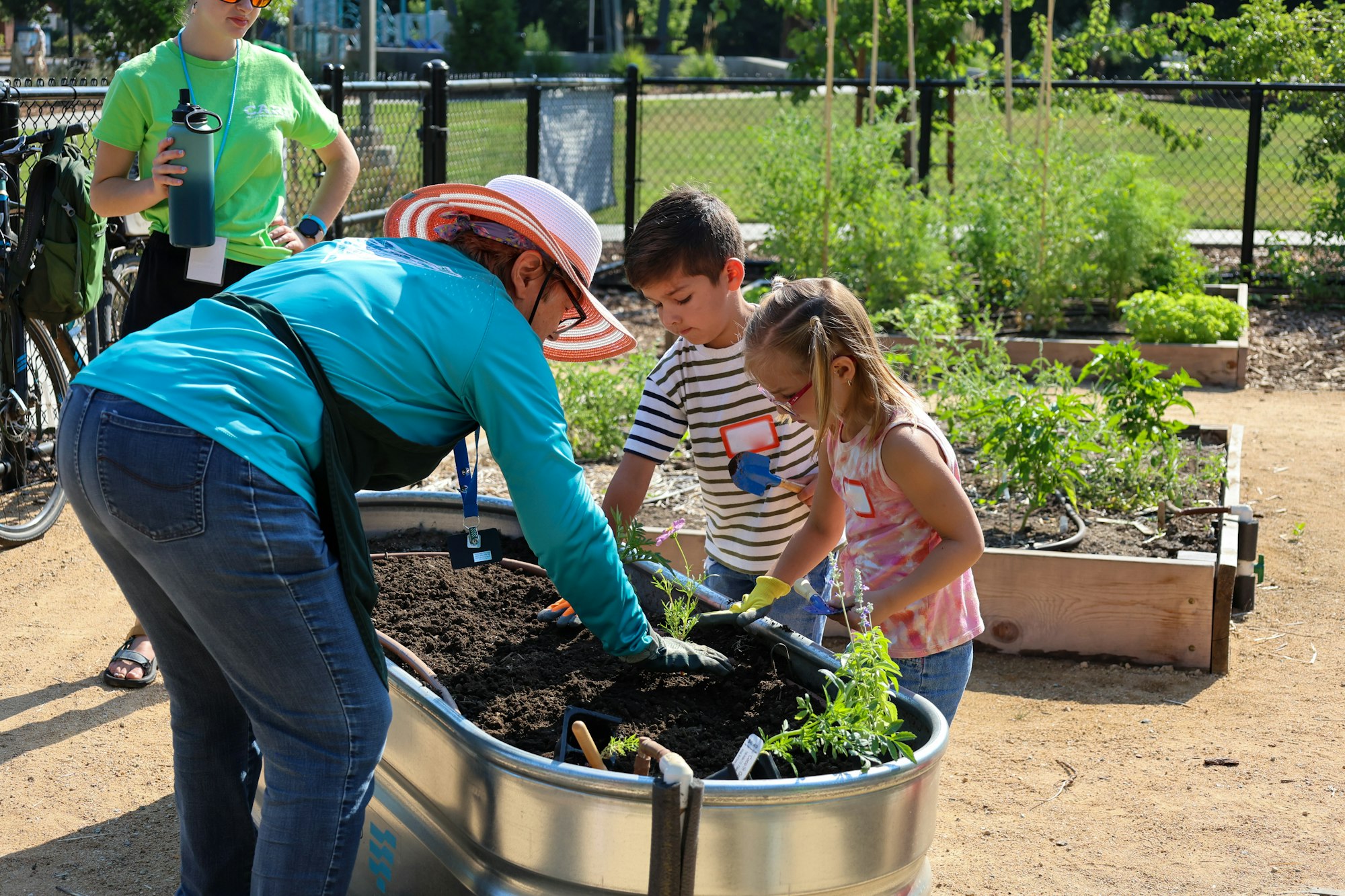 A woman and two children are gardening in a community garden, planting in a metal container filled with soil.