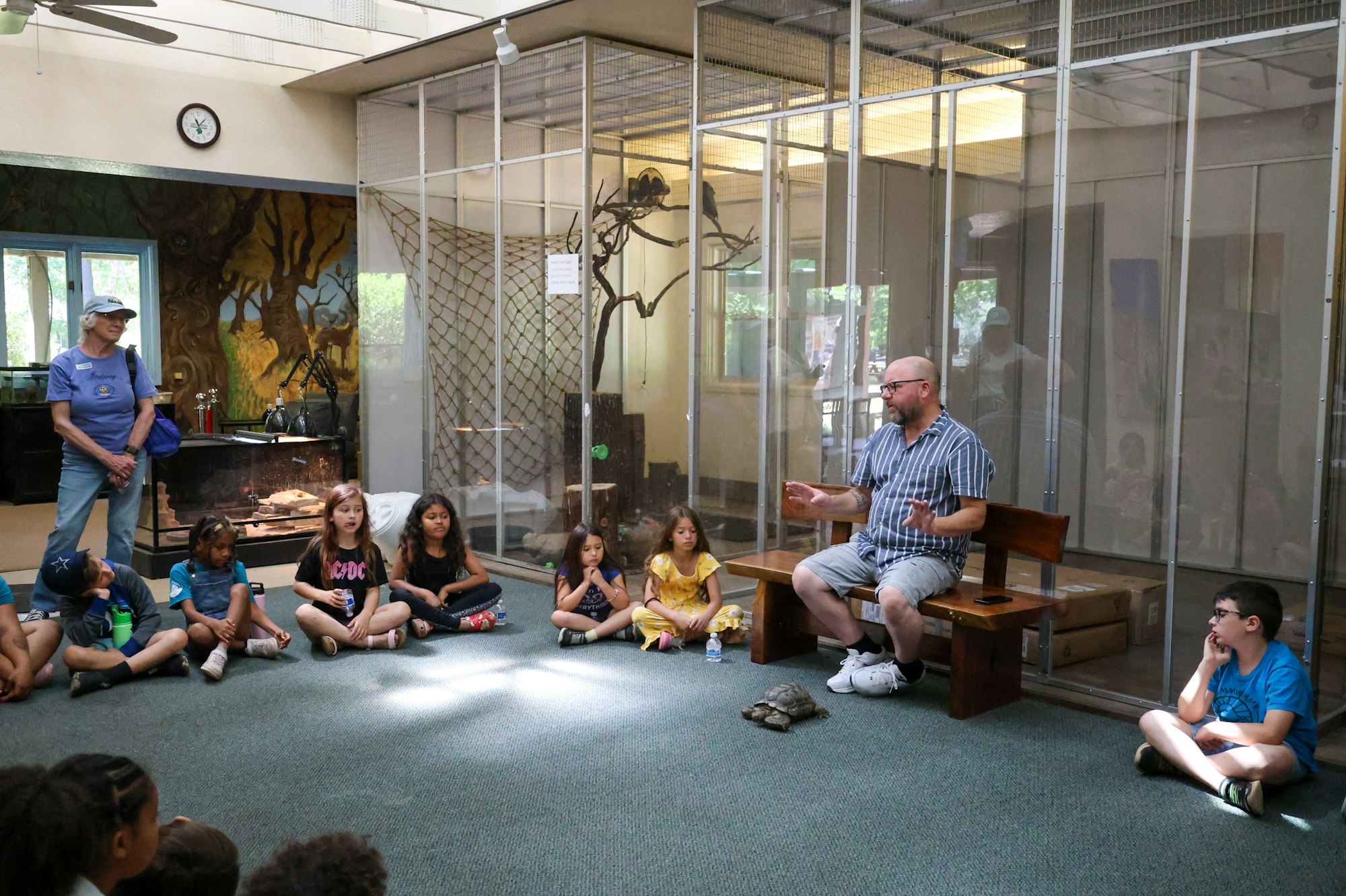 A group of children sitting on a floor, listening to a man with a tortoise nearby, in an indoor space with cages and mural art.