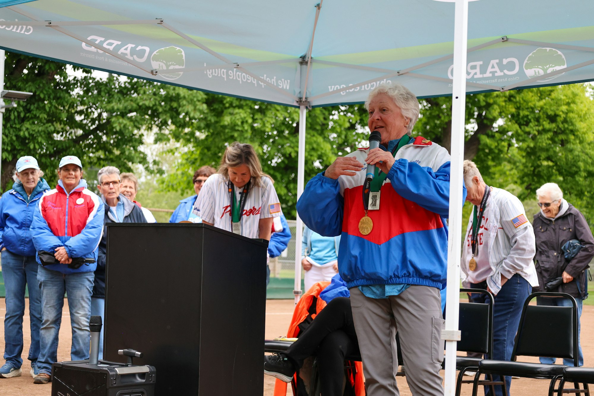 A person speaks into a microphone under a canopy, wearing a medal, with others standing nearby.