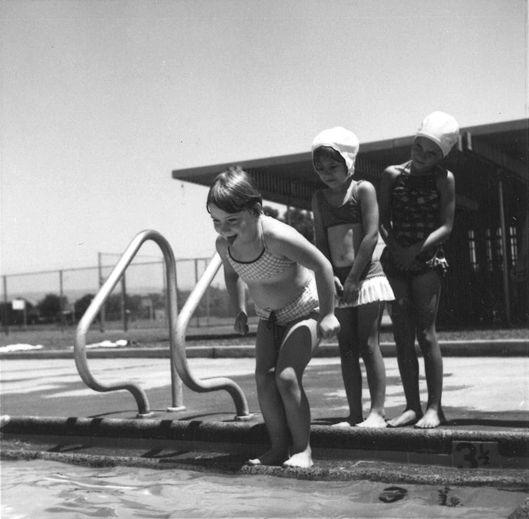 Three children in swimsuits at a poolside; the first child is preparing to jump in, others watching.