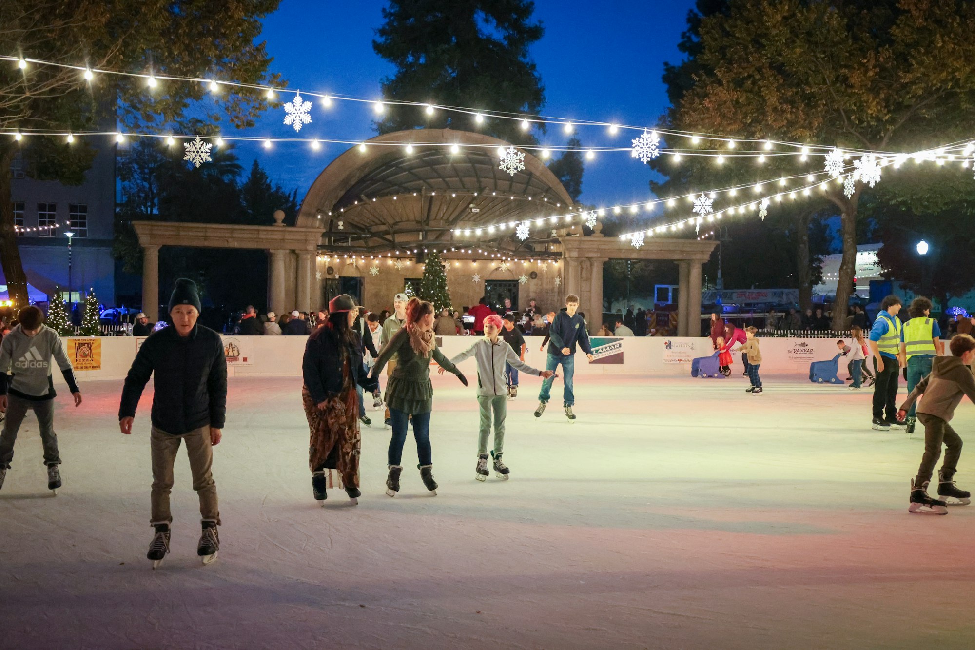 People ice skating at a festive outdoor rink decorated with lights and snowflakes at dusk.