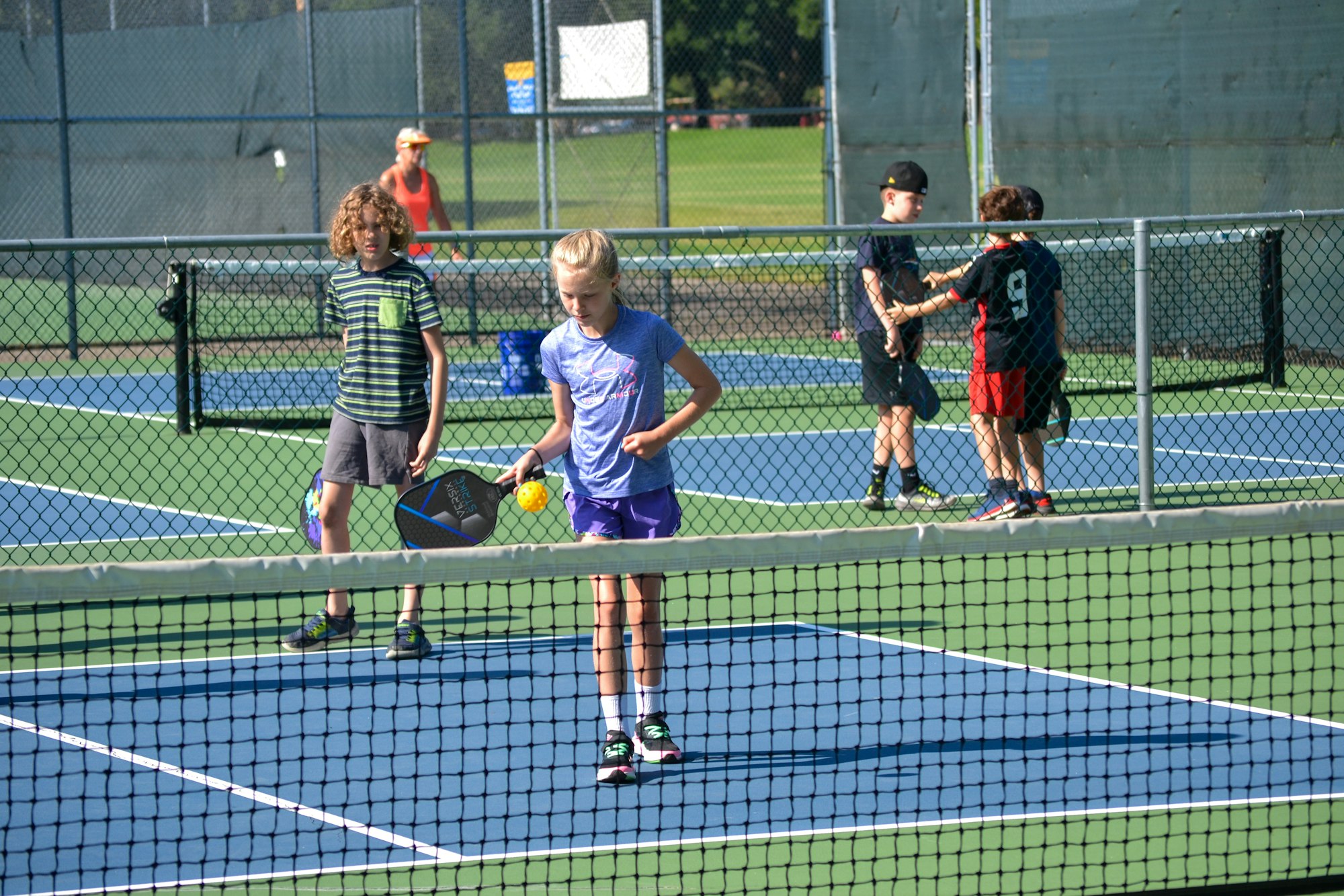 Kids playing pickleball on an outdoor court with paddles and a yellow ball.
