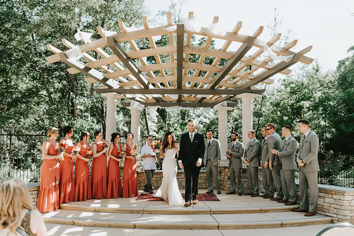 A wedding ceremony with a couple at the altar, bridesmaids in red, and groomsmen in gray suits, under a pergola.
