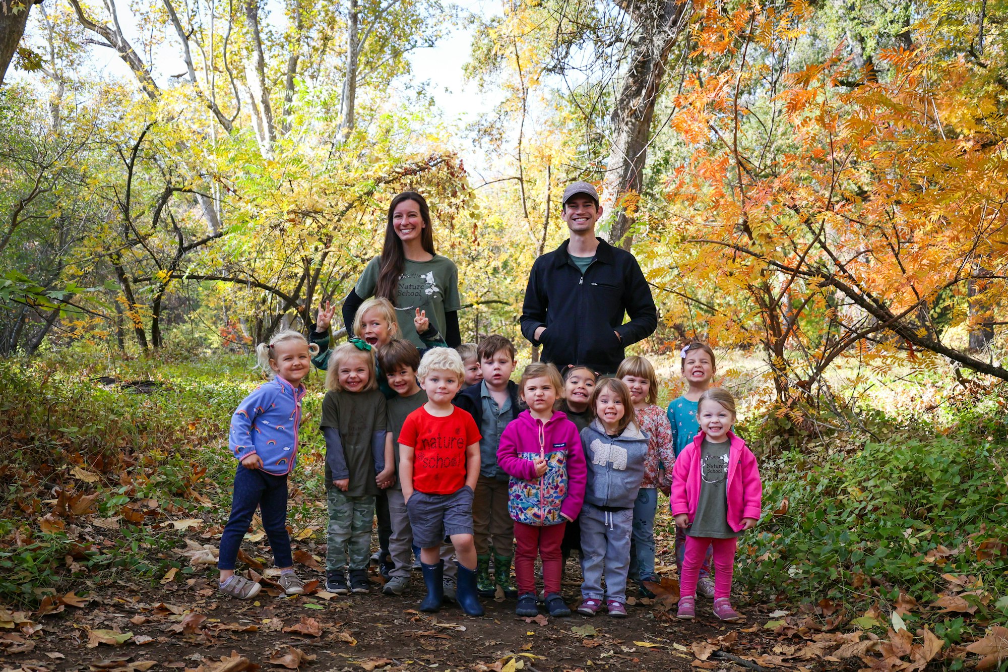 A group of children and two adults smiling in an autumn forest setting.