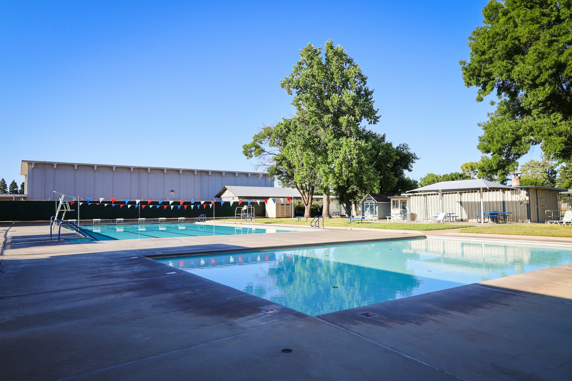 Outdoor swimming pool with diving board, colorful flags, trees, and buildings in the background under a clear blue sky.