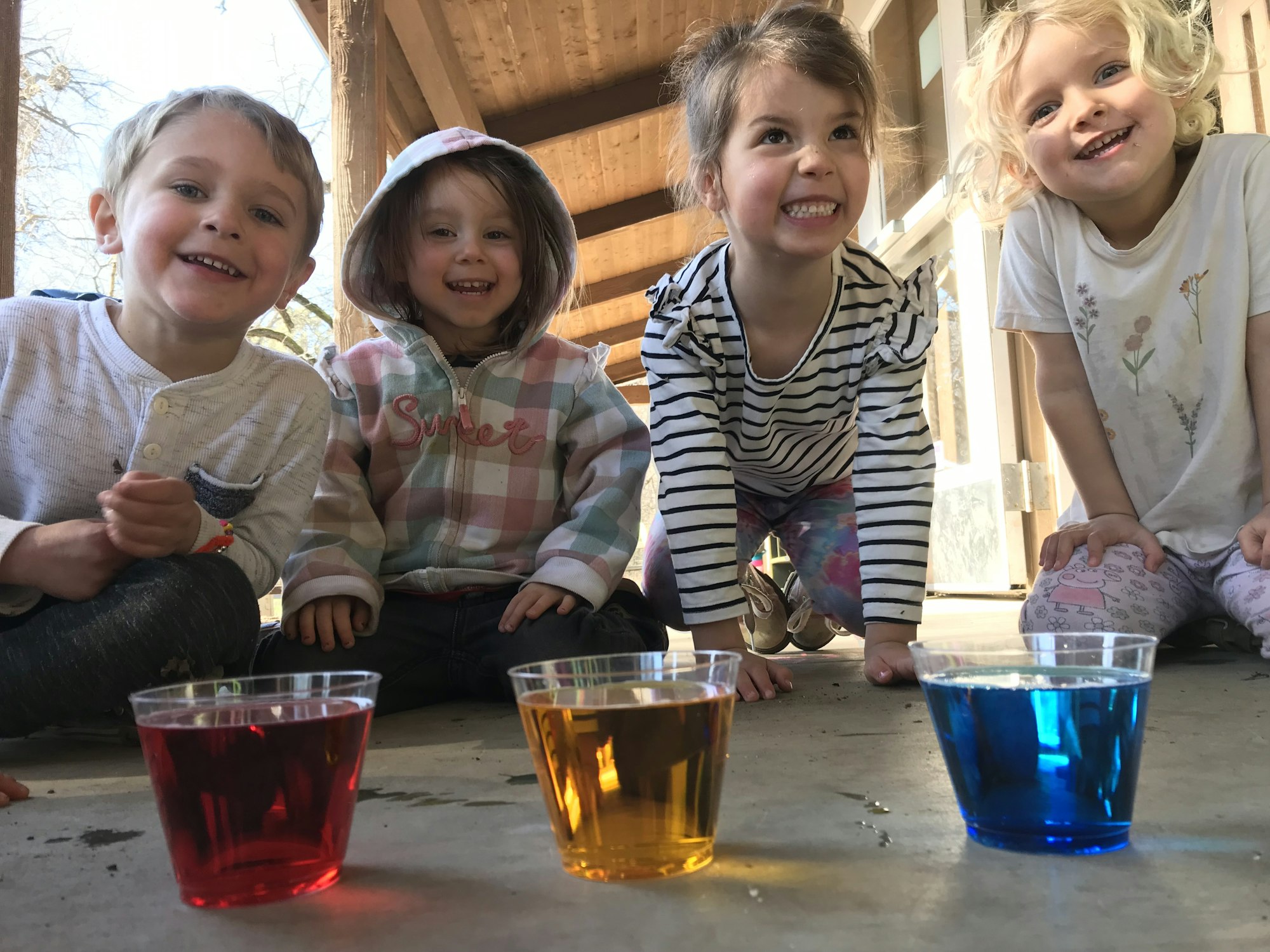 Four kids smiling next to cups filled with red, yellow, and blue liquid.