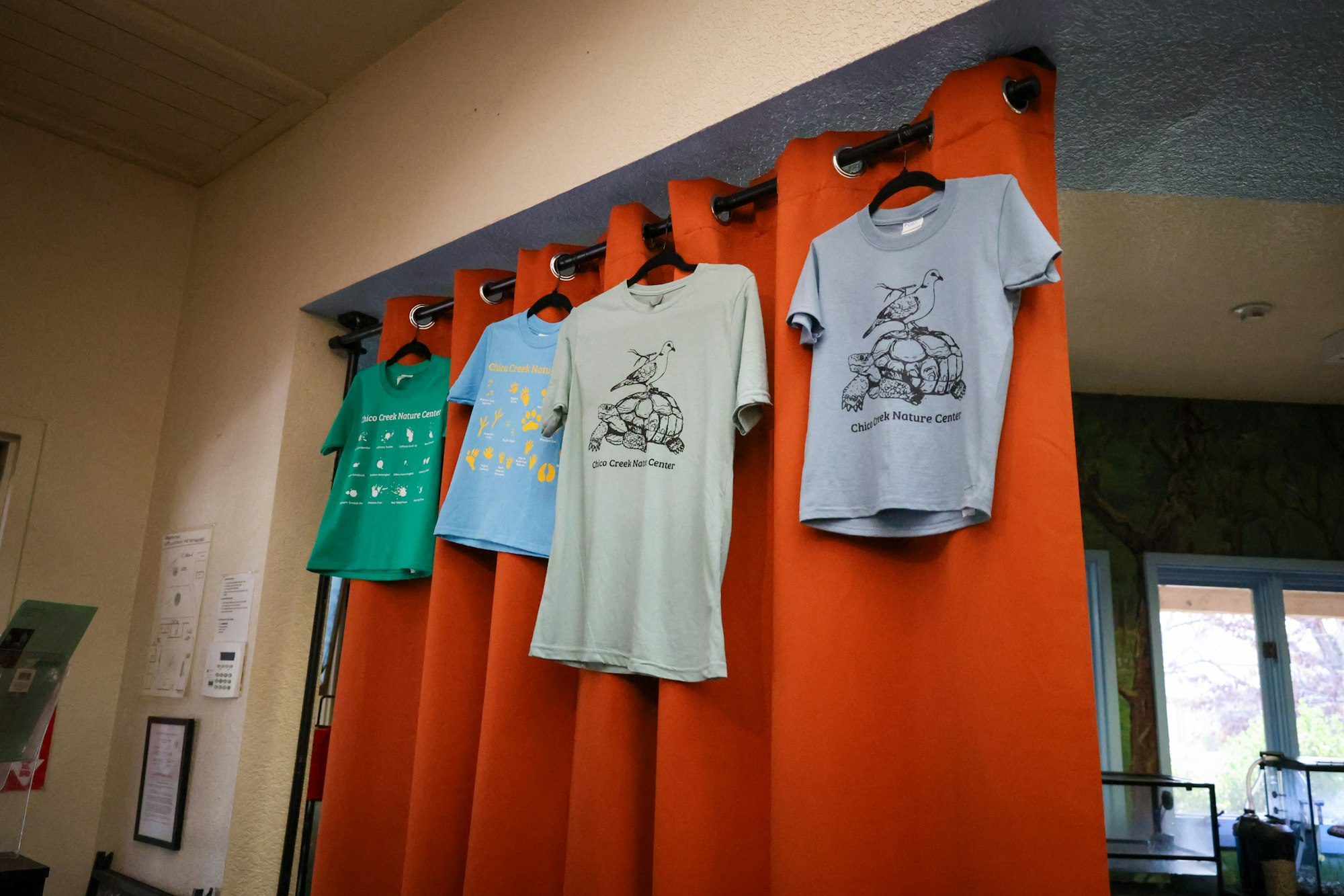 The image shows several colorful t-shirts hanging on an orange curtain, likely promoting the Chico Creek Nature Center.