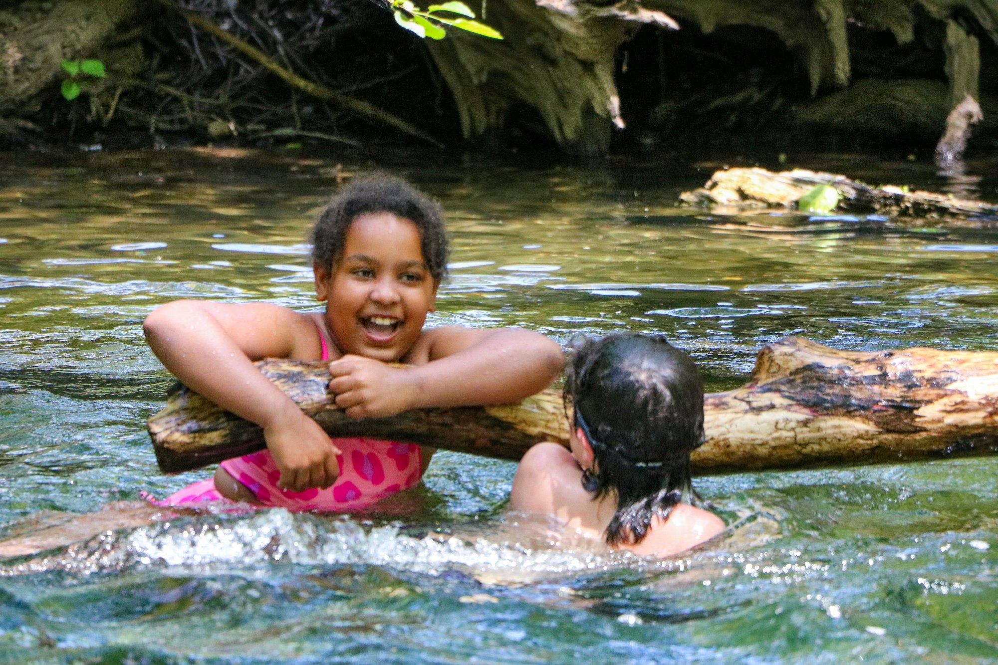 Two kids playing in a river, one smiling at the camera, holding onto a log.