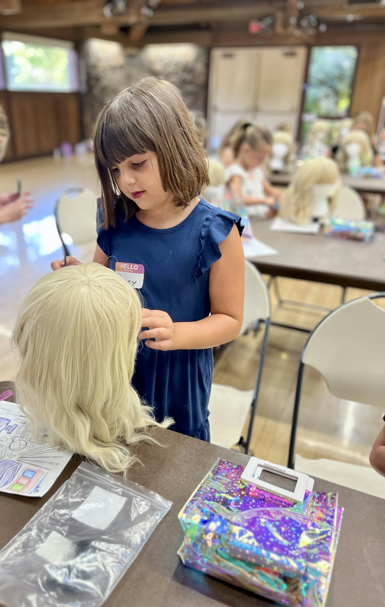 A girl styles a wig at a table, surrounded by other children working on similar projects in a bright indoor space.