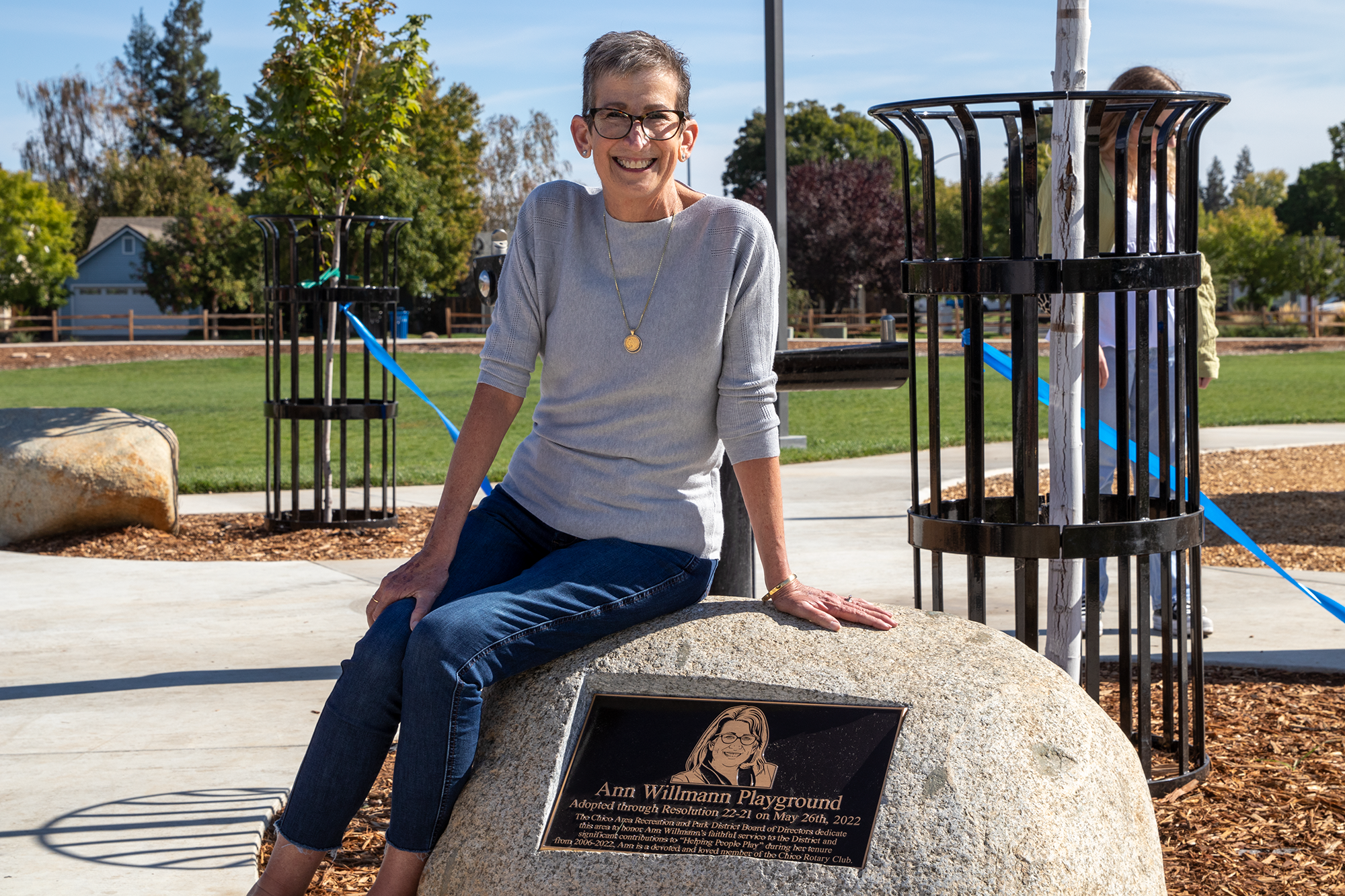 A person sitting on a rock with a playground plaque, smiling in a park setting.