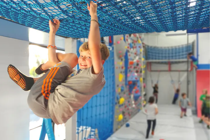 Child hanging from a blue net in an indoor play area with climbing structures and other kids in the background.