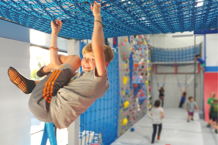 Child hanging from a blue net in an indoor play area with climbing structures and other kids in the background.