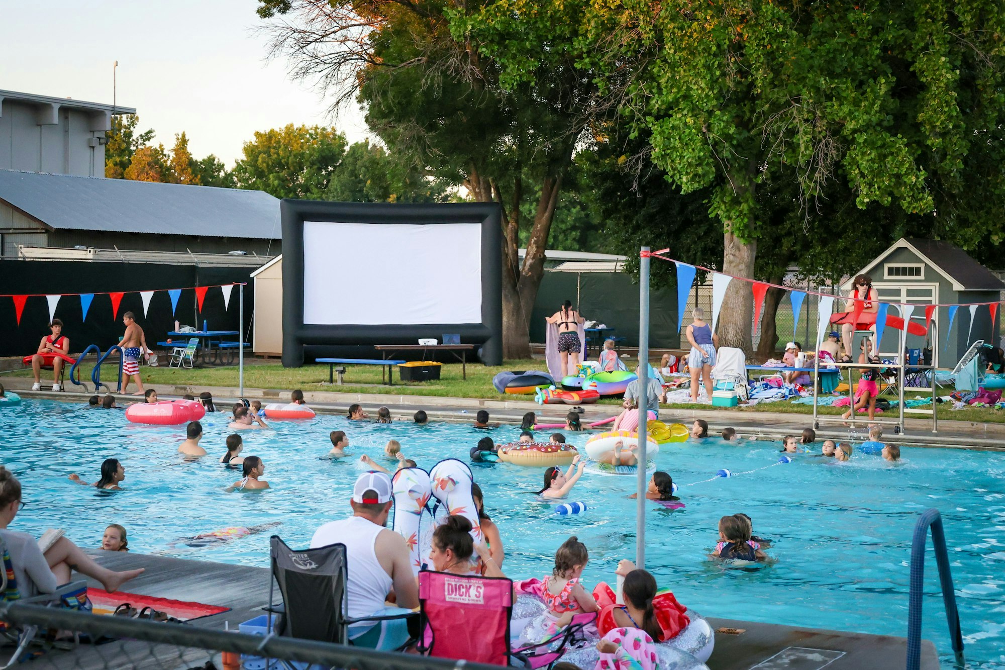 A lively pool scene with people swimming, floaties, and an inflatable screen for a movie night. Fun summer vibes!