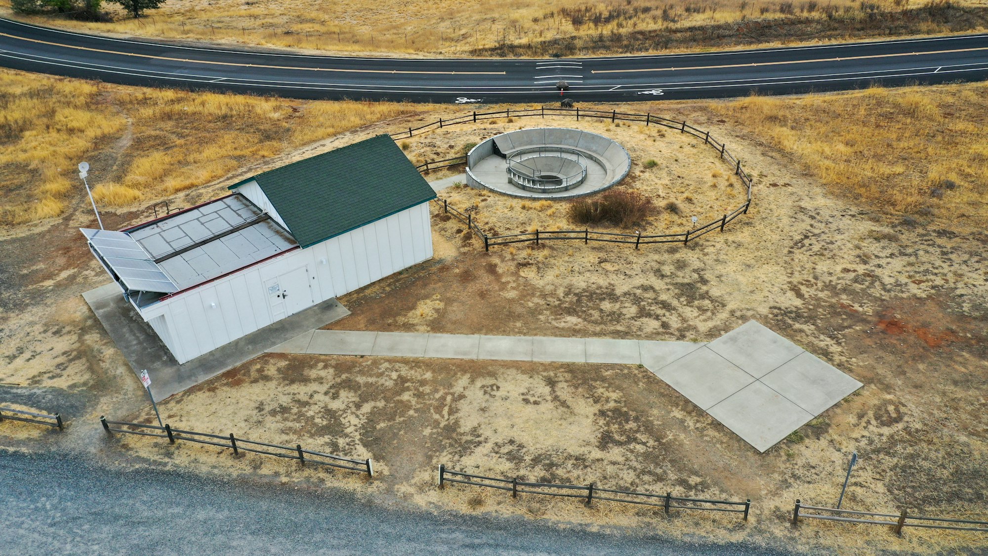 Aerial view of a building with solar panels, a circular fenced structure nearby, and a road in the background.