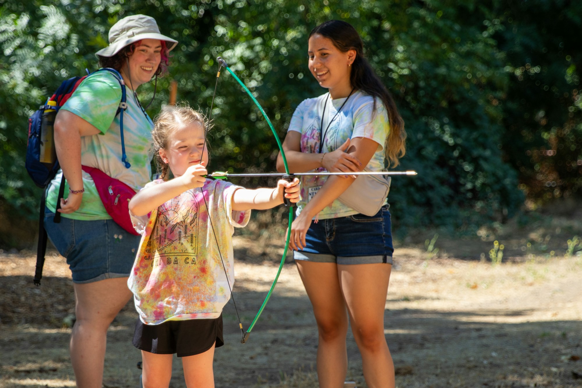 A child practices archery with guidance from two smiling adults outdoors.