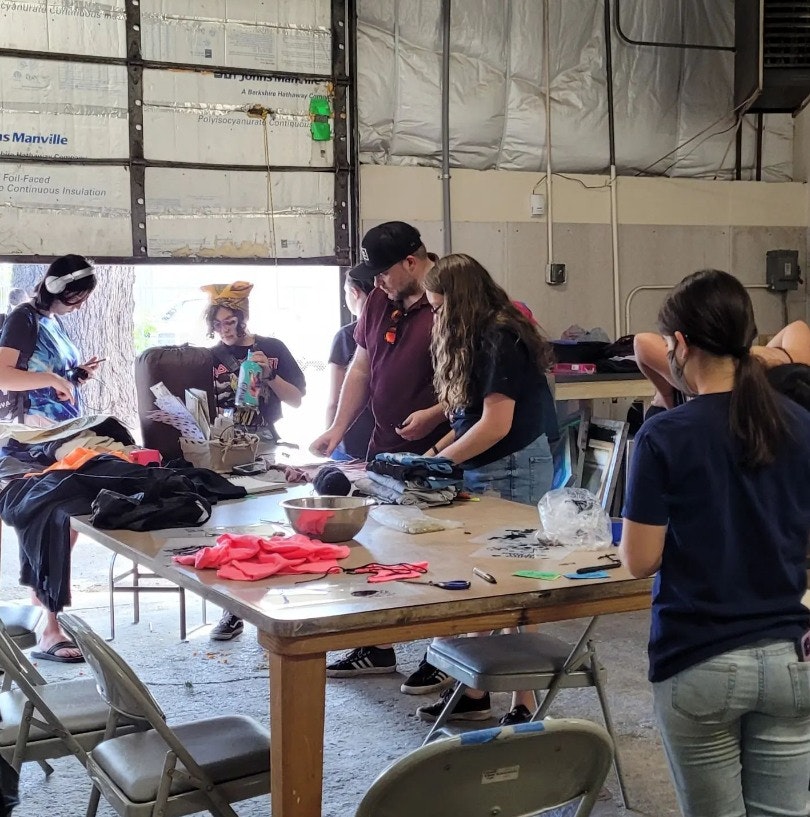 A group of people in a workshop collaboration, creating or organizing materials around a table filled with clothing and supplies.
