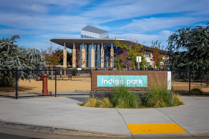 A sign for Indigo Park is shown, featuring modern architecture, greenery, and a fenced area with a playground structure.