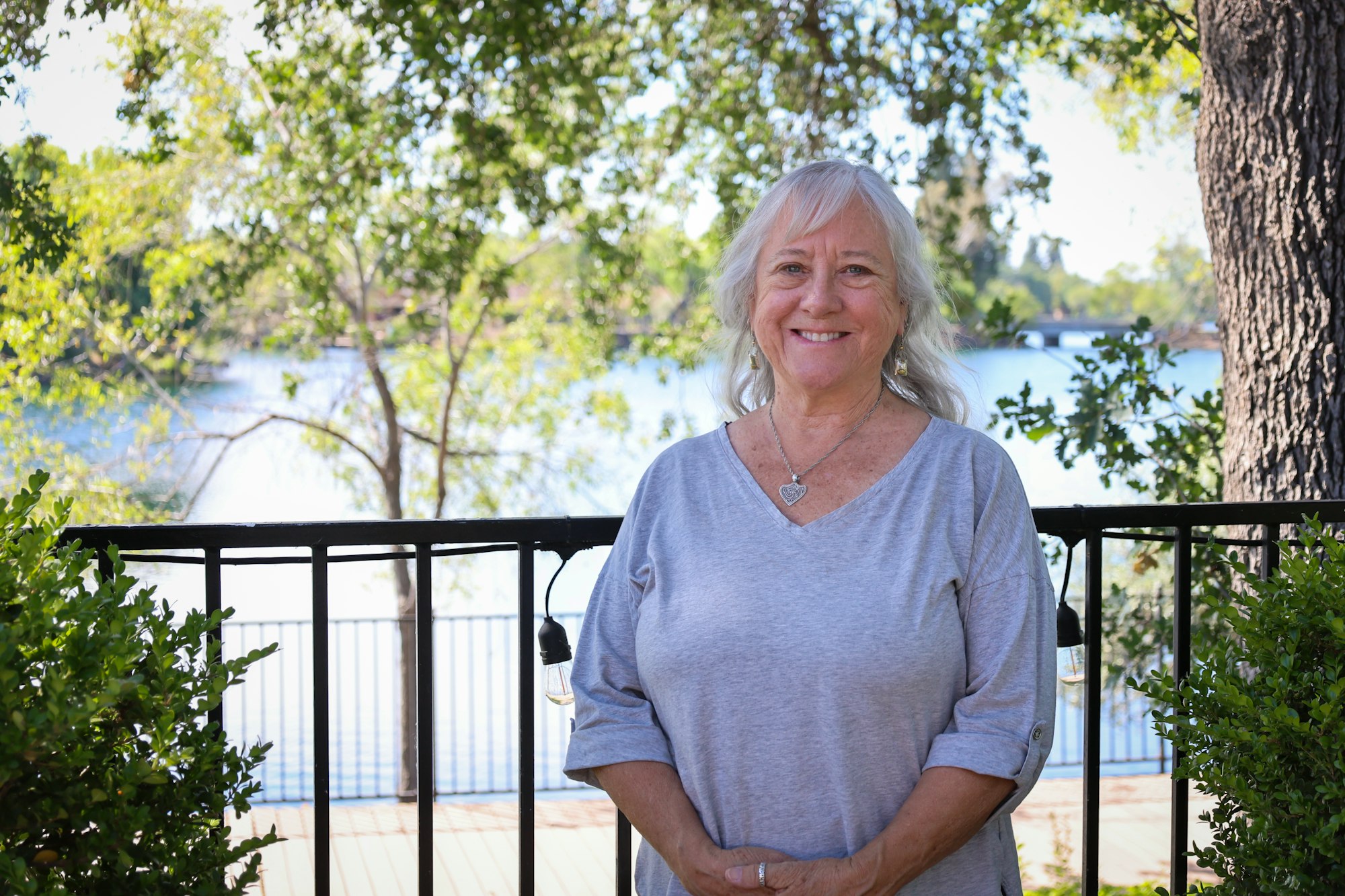 A smiling woman standing by a railing with a lake and trees in the background.