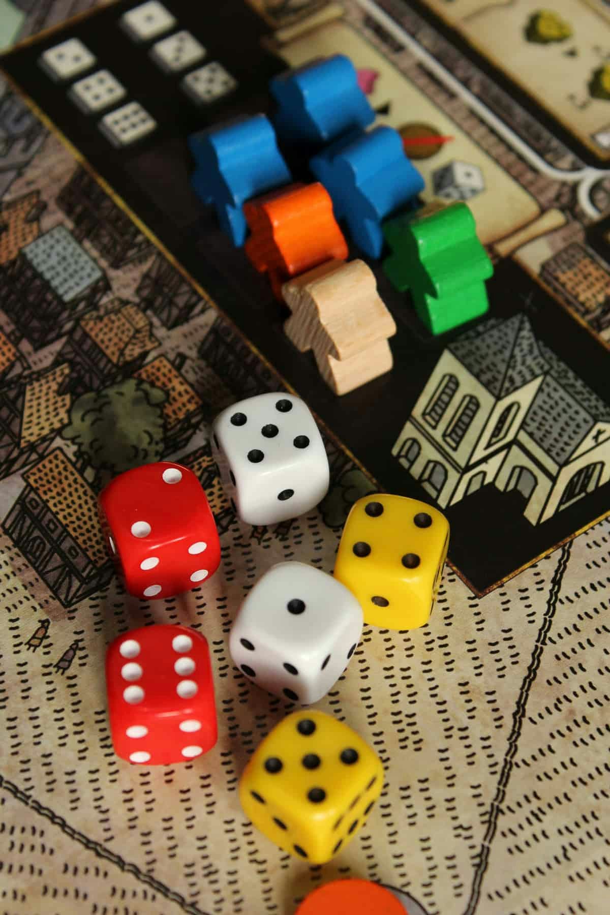 The image features colorful dice and wooden game pieces on a board game surface, suggesting a strategy or board game setup.