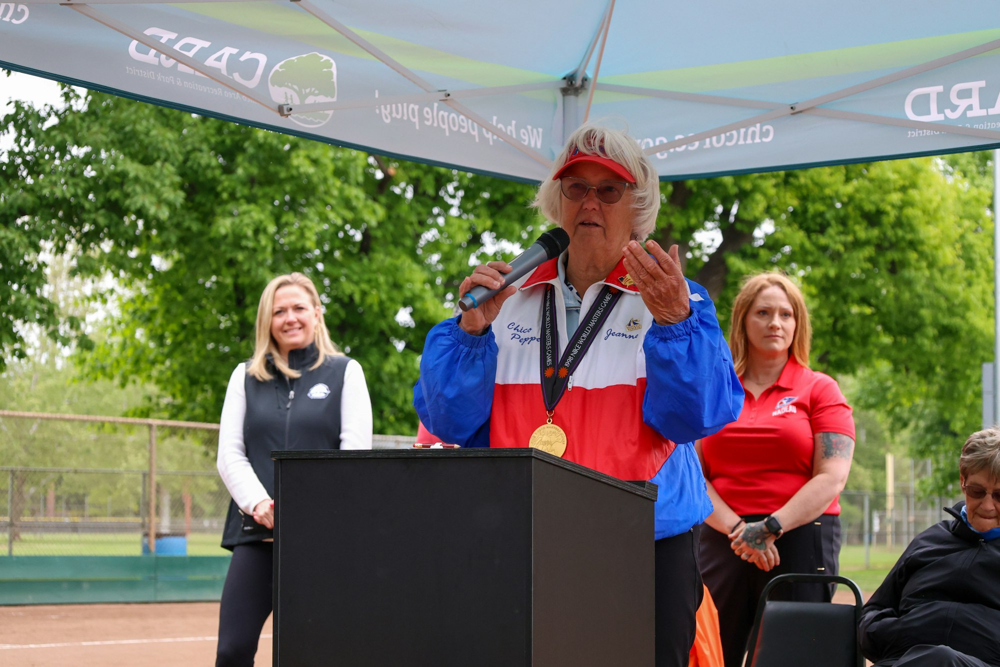 A person speaking at a podium under a canopy, wearing a jacket with a medal. Others stand nearby outdoors.