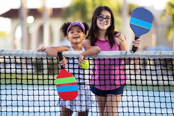 Two girls are smiling and posing by a net, holding paddles and a ball, ready to play a game, likely pickleball or similar.