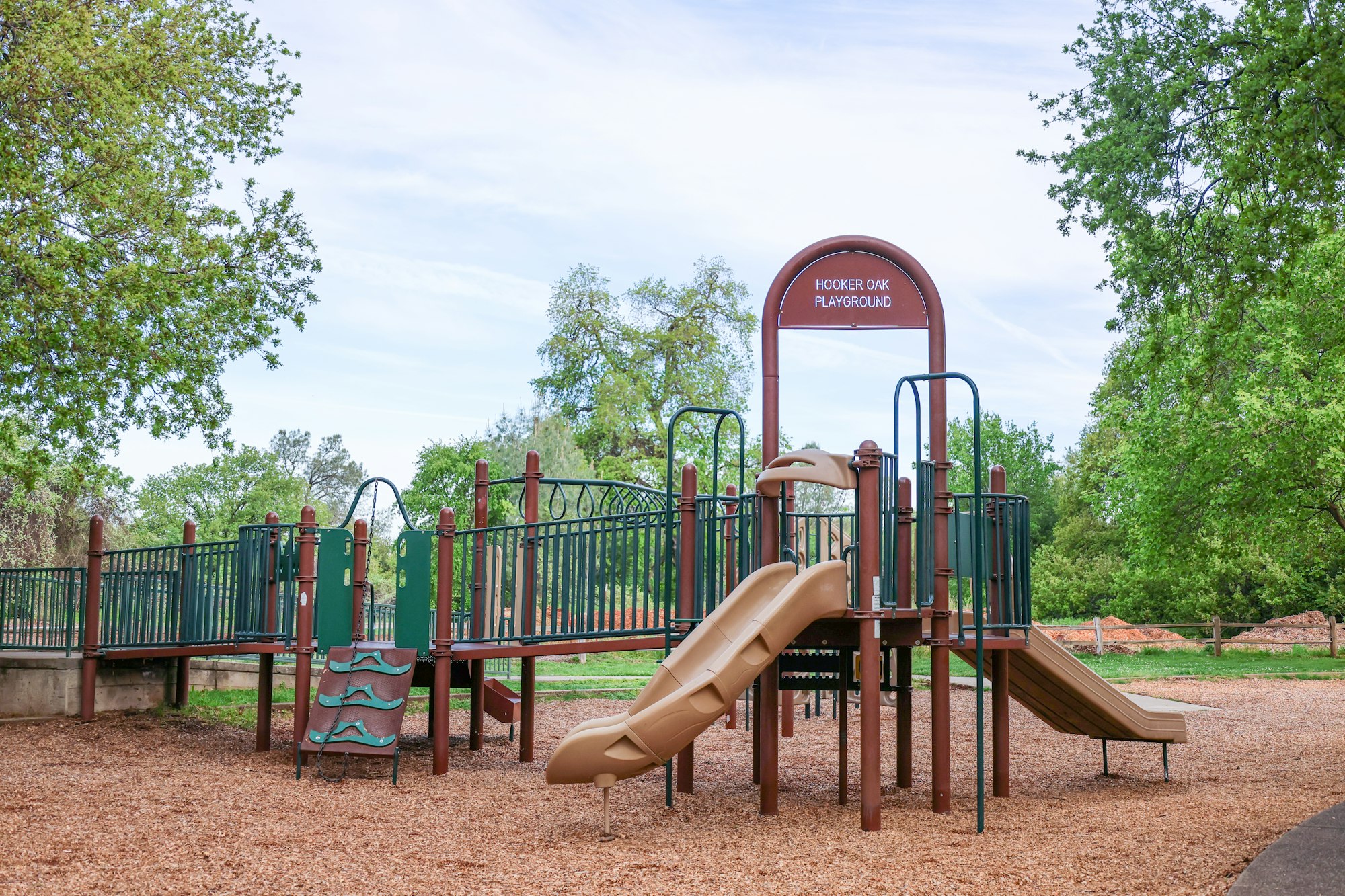 A playground with slides and climbing structures on a sunny day, surrounded by trees.