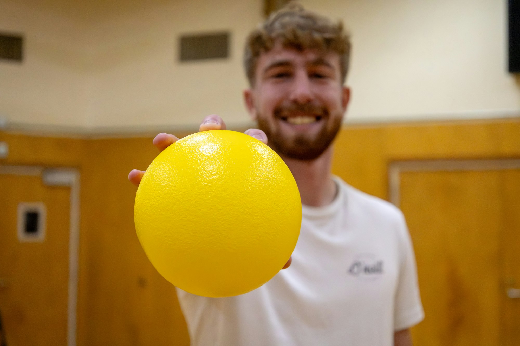 A smiling person holds a bright yellow ball in a gym-like setting, ready for a game or activity.