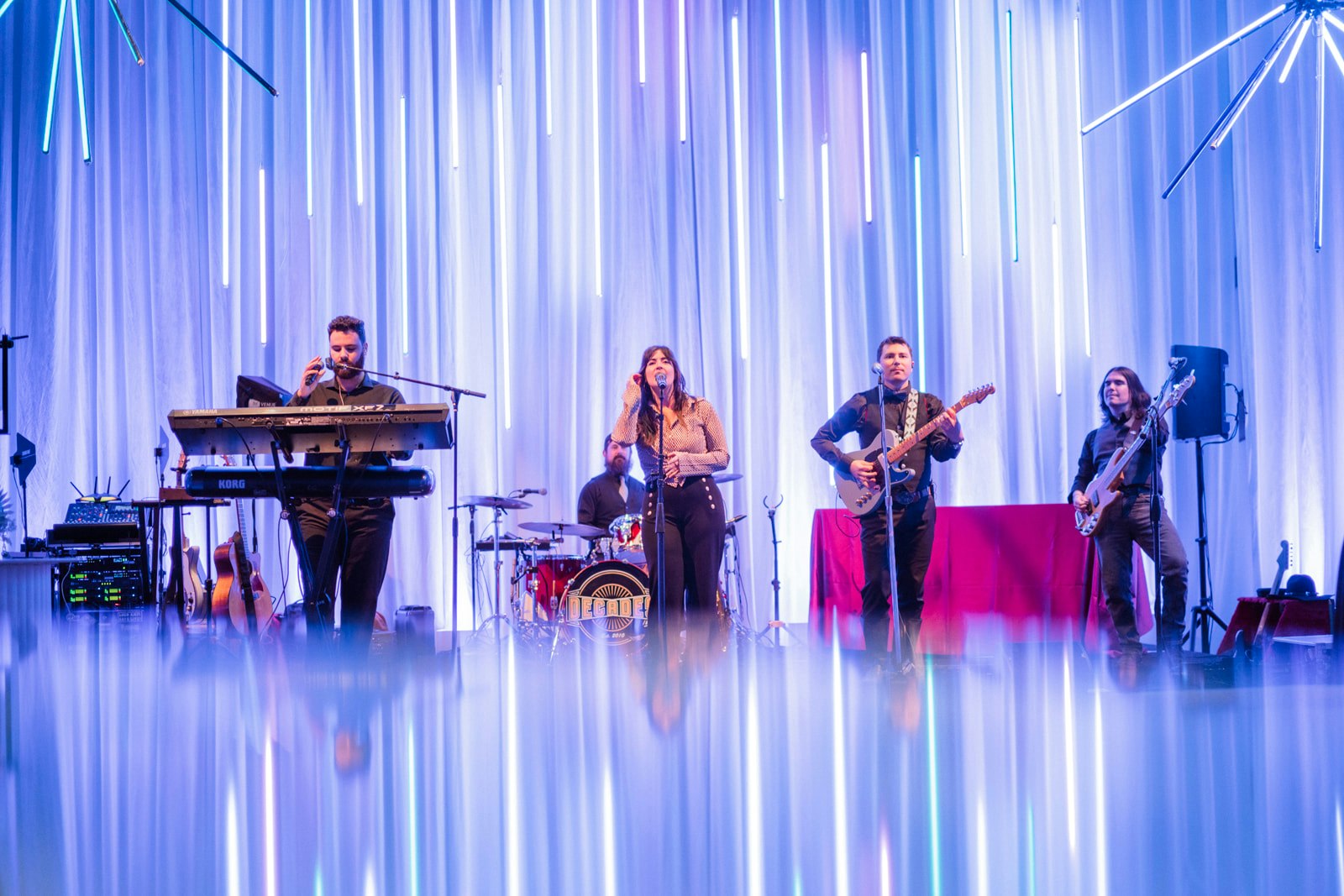 A band performing on stage with keyboards, drums, guitar, and bass under blue vertical lights and reflections on the floor.