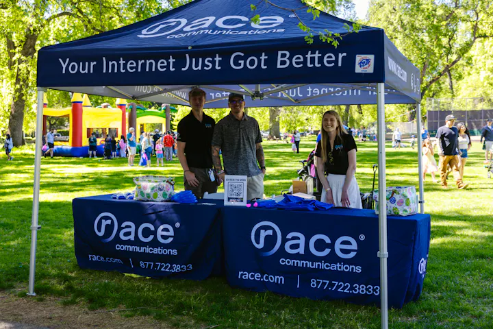 Outdoor race communications booth with three people under a tent, offering promotions.