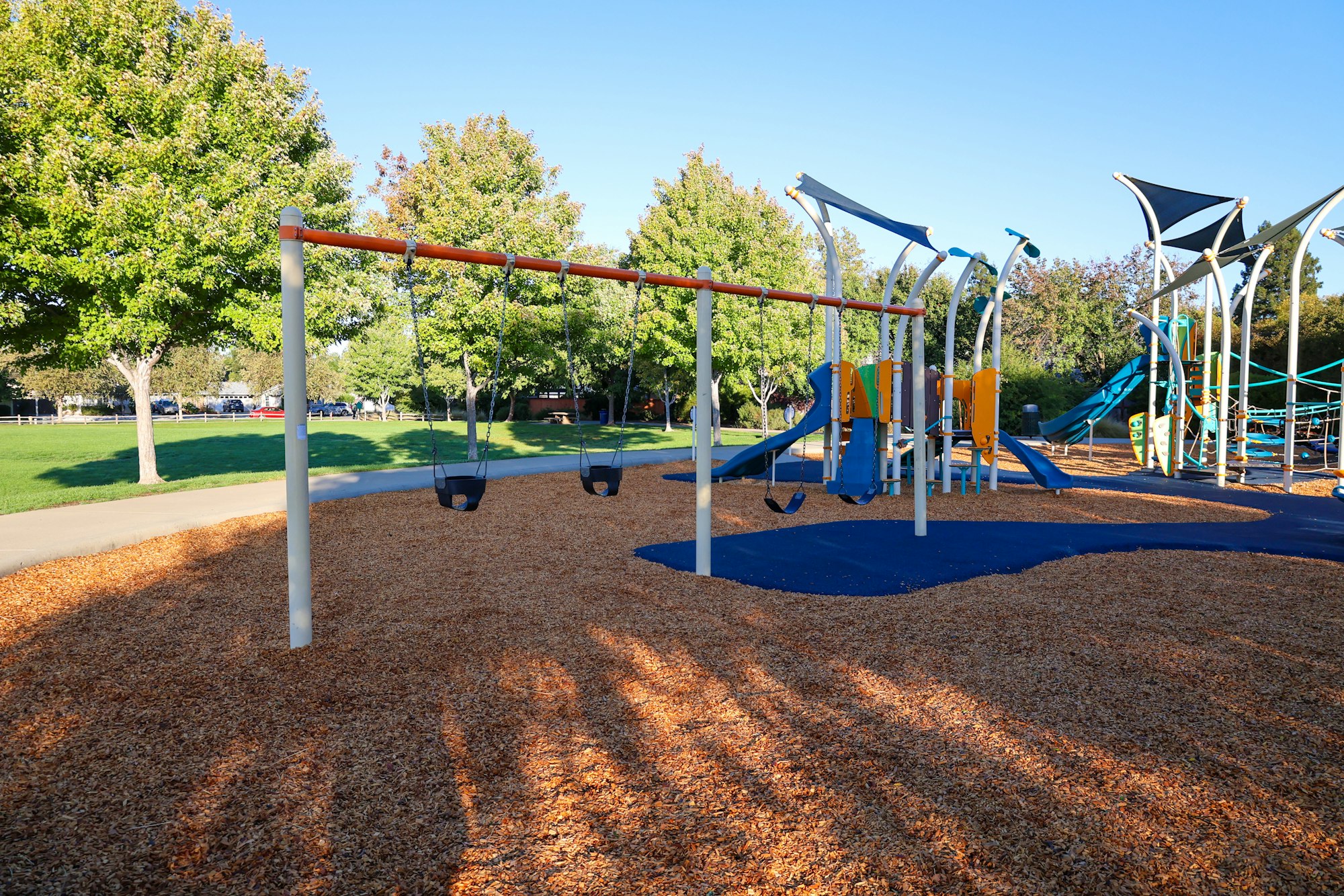 A sunny playground featuring swings, slides, and play equipment surrounded by trees and wood chip ground cover.