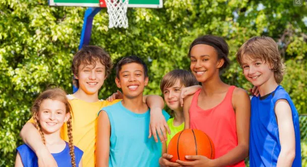 A group of six diverse kids poses together with a basketball, smiling in front of a basketball hoop on a sunny day.