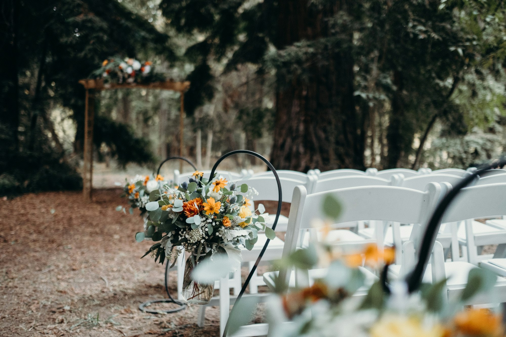 Outdoor wedding setup with white chairs and floral decorations in a forest setting.