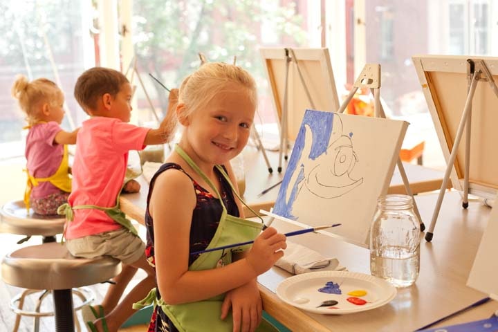 Children painting at easels in a bright room, wearing aprons. A girl smiles at the camera, holding a brush with paint.